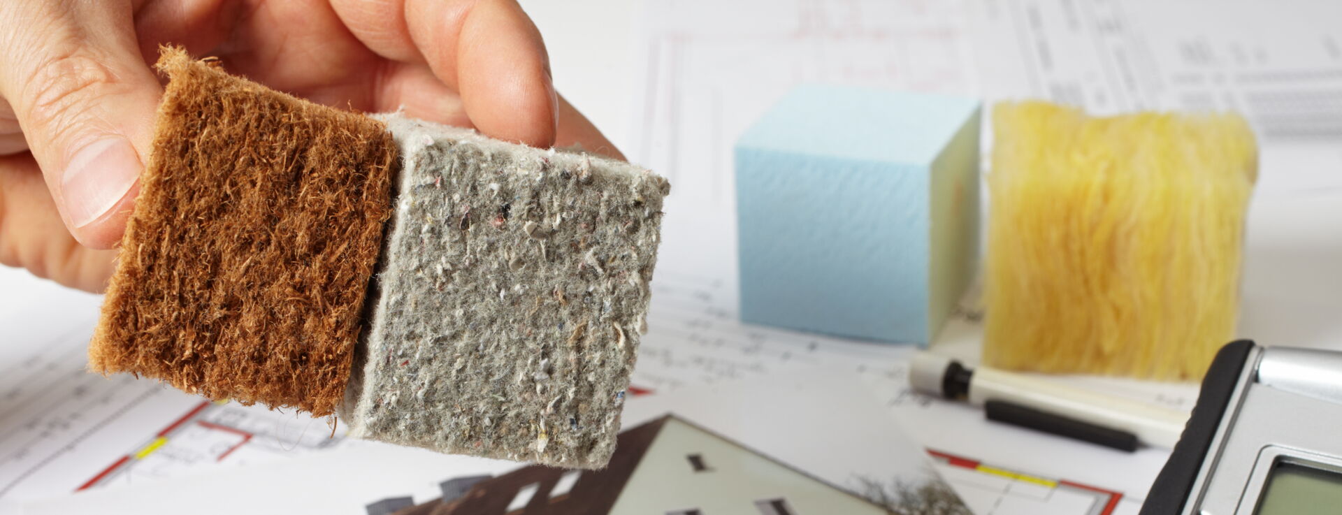 Hand hält Dämmstoffmuster über Thermografiebild eines Hauses, Taschenrechner und Pläne im Hintergrund. Energieberatung in Südsachsen.